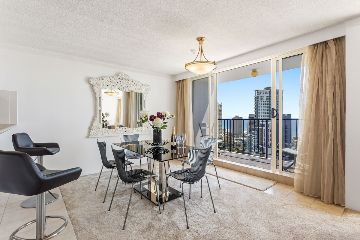 A modern dining area with a glass table and chairs, a decorative mirror, and a view of the cityscape through large windows.