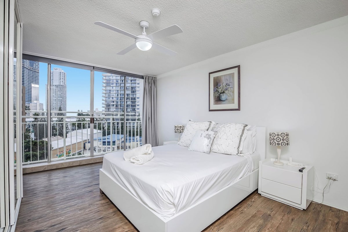 A modern bedroom with a large bed, a ceiling fan, a nightstand with a lamp, and a city view through large windows.