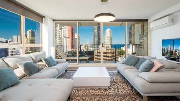 Amplio salón con un ventanal que ofrece vistas a la ciudad, una mesa de centro blanca y dos sofás con cojines.