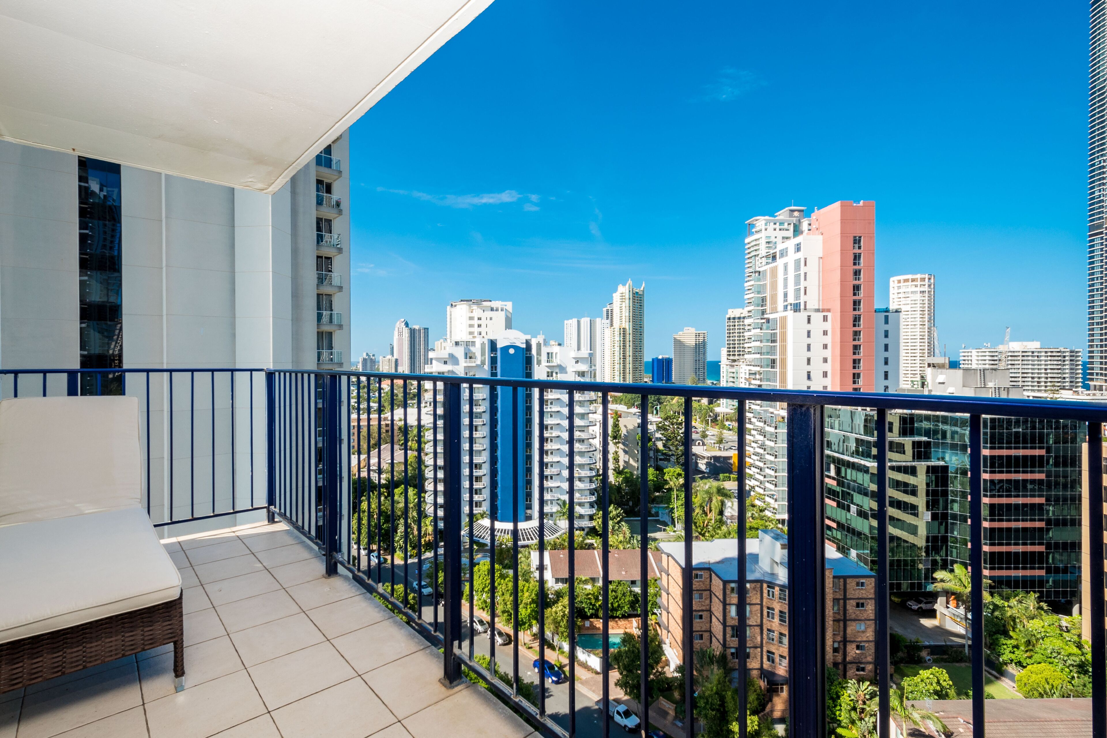 A balcony with a white bench, a view of a cityscape with various buildings, and a clear blue sky.