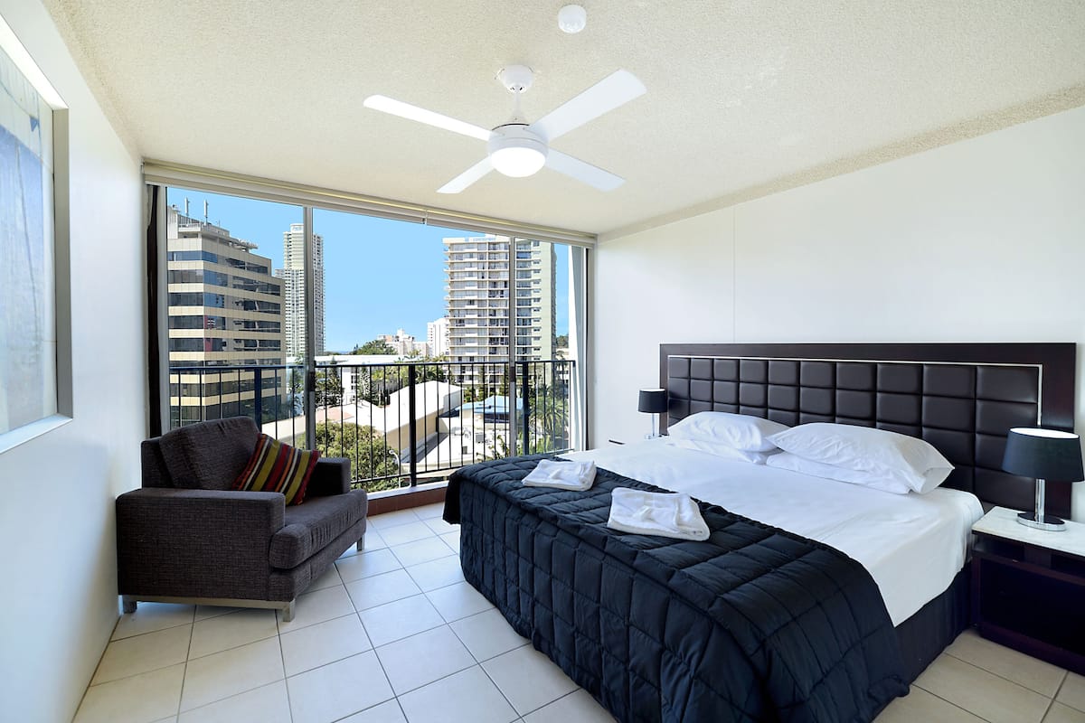 A modern hotel room with a large bed, a sofa, a ceiling fan, and a balcony with a city view.