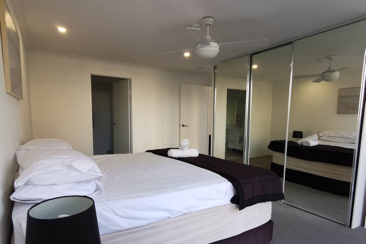 A modern hotel room with a large bed, a bedside lamp, a ceiling fan, and a mirror wall.