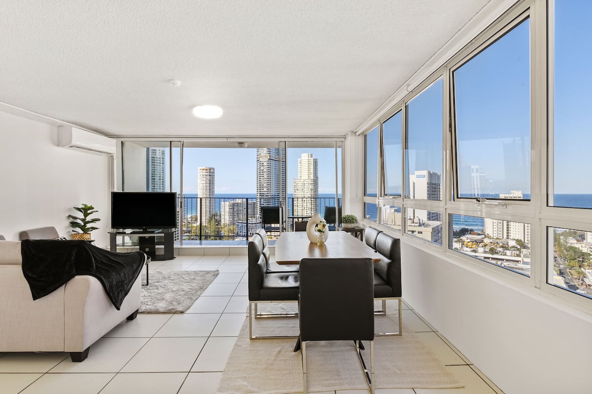 A modern living room with a large window, a sofa, a TV, and a dining area.