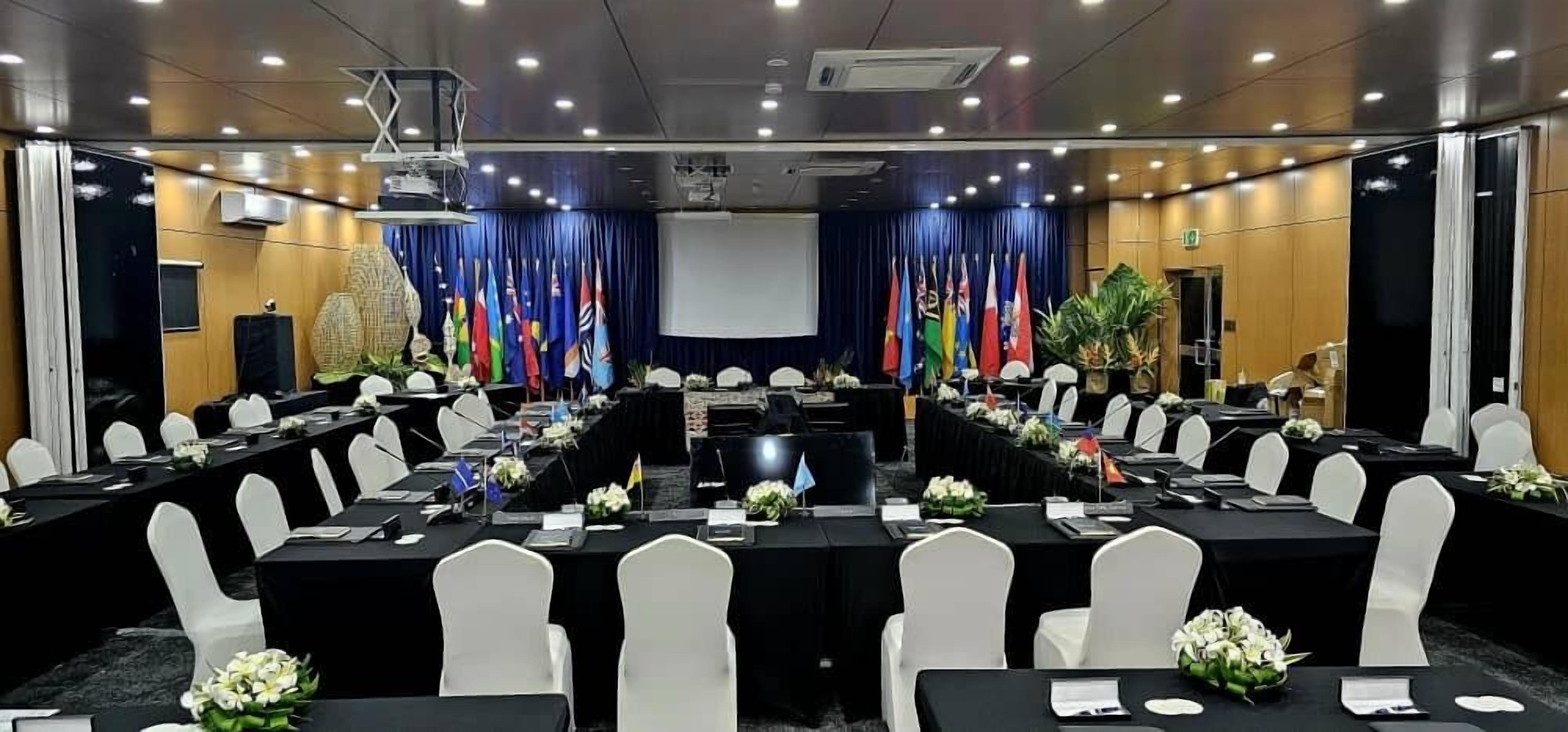 Un salón de conferencias con varias mesas, sillas blancas y un escenario con banderas.