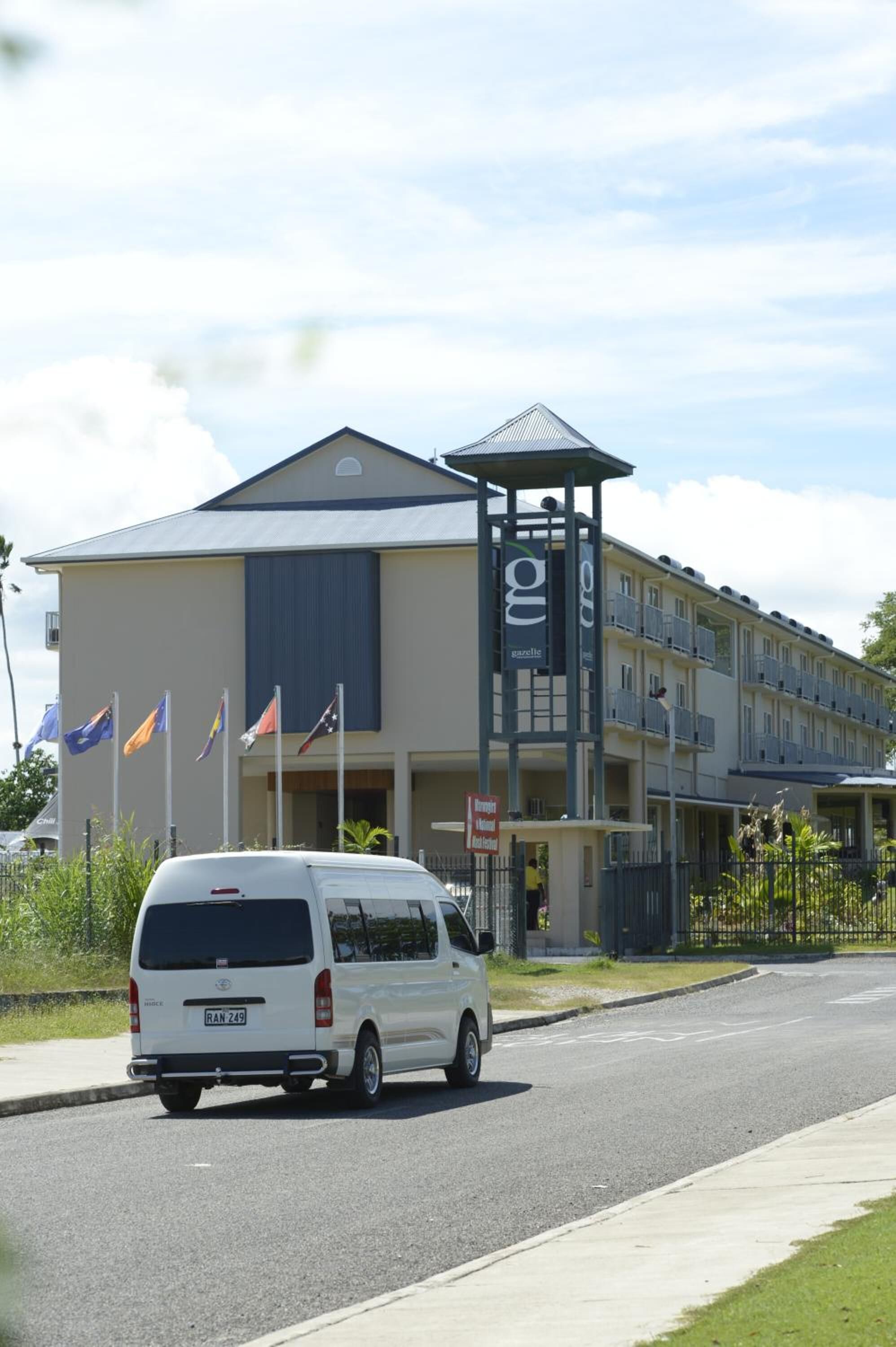 A white van parked in front of a building with multiple flags, a covered entrance, and a balcony area.