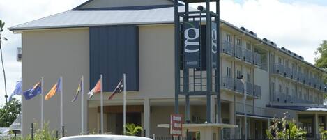 A white van parked in front of a building with multiple flags, a covered entrance, and a balcony area.