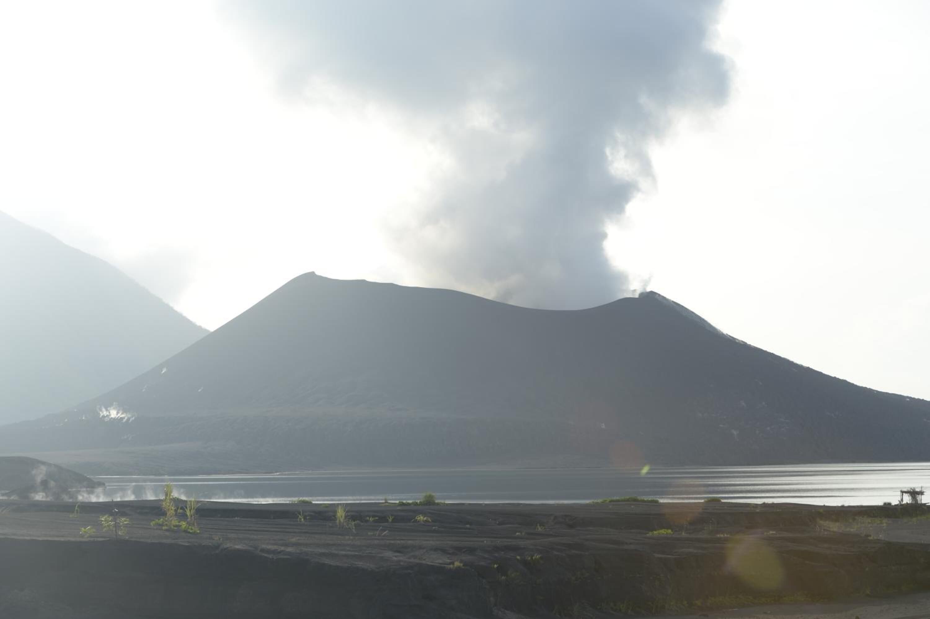 Ein Berg mit einer Rauchwolke, ein Gewässer und eine karge Landschaft.
