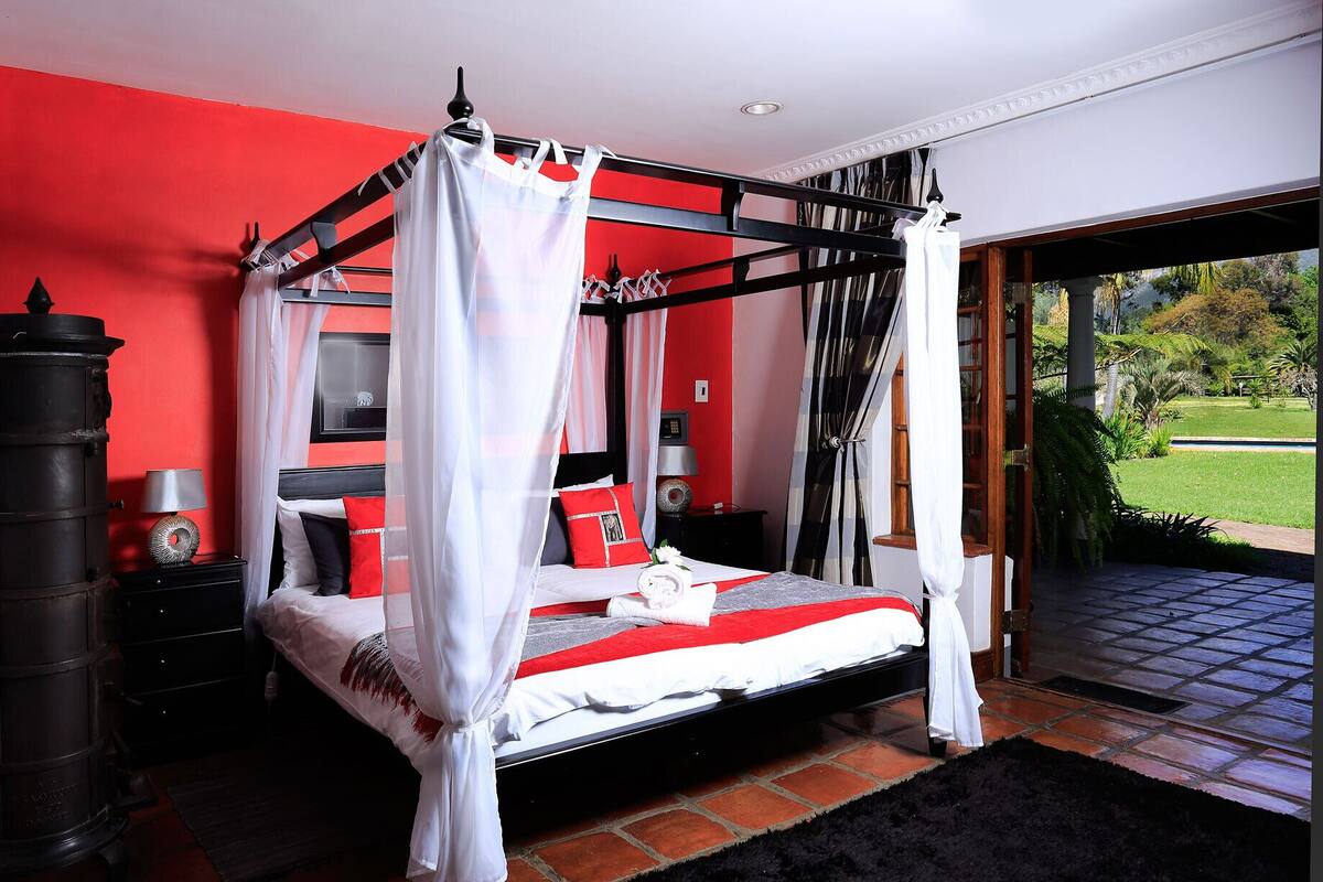 Un lit à baldaquin, un radiateur noir, une table de chevet avec une lampe et une vue sur un jardin par une porte vitrée.