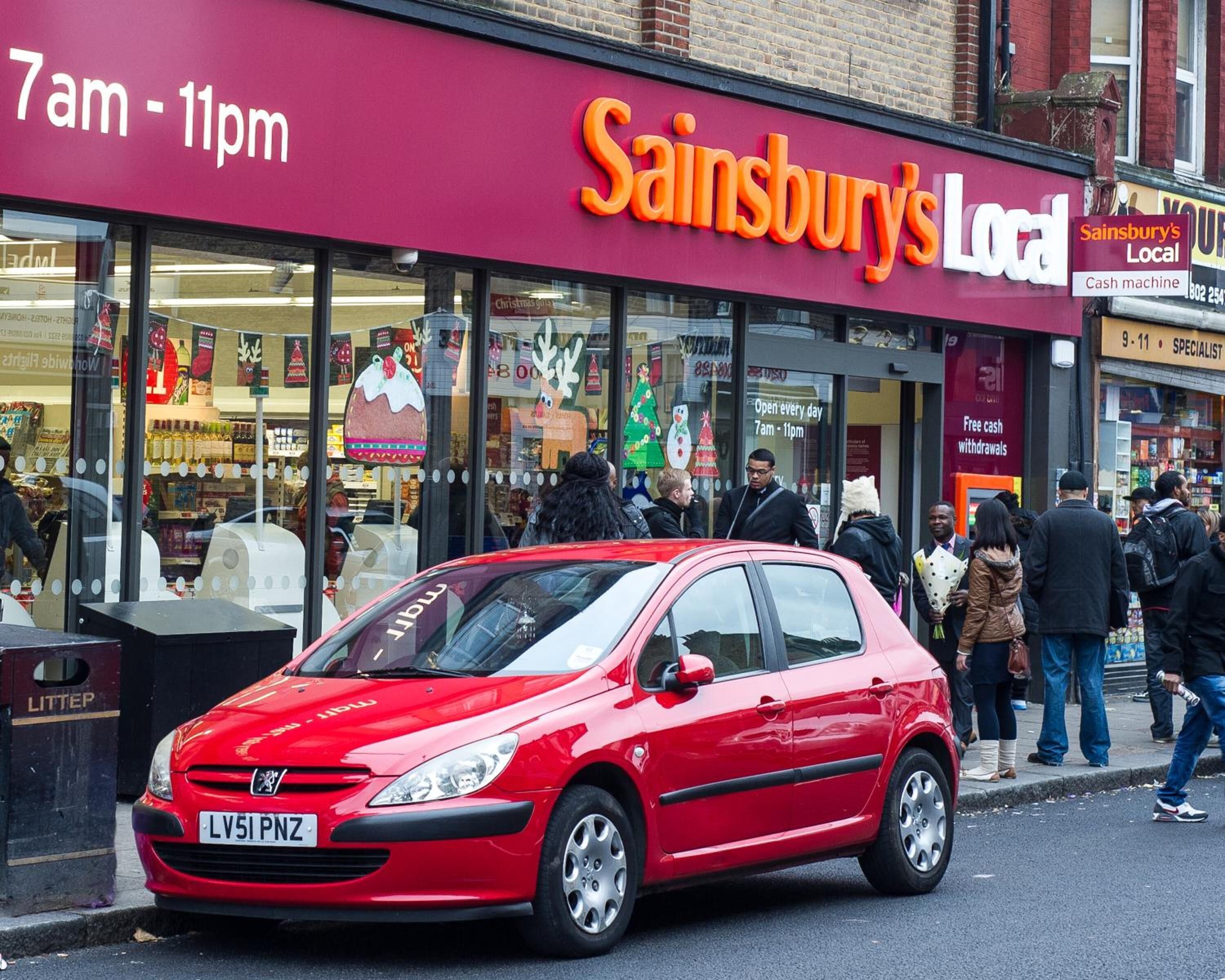 Un coche rojo aparcado frente a una tienda Sainsbury's Local, con escaparate visible y gente afuera.