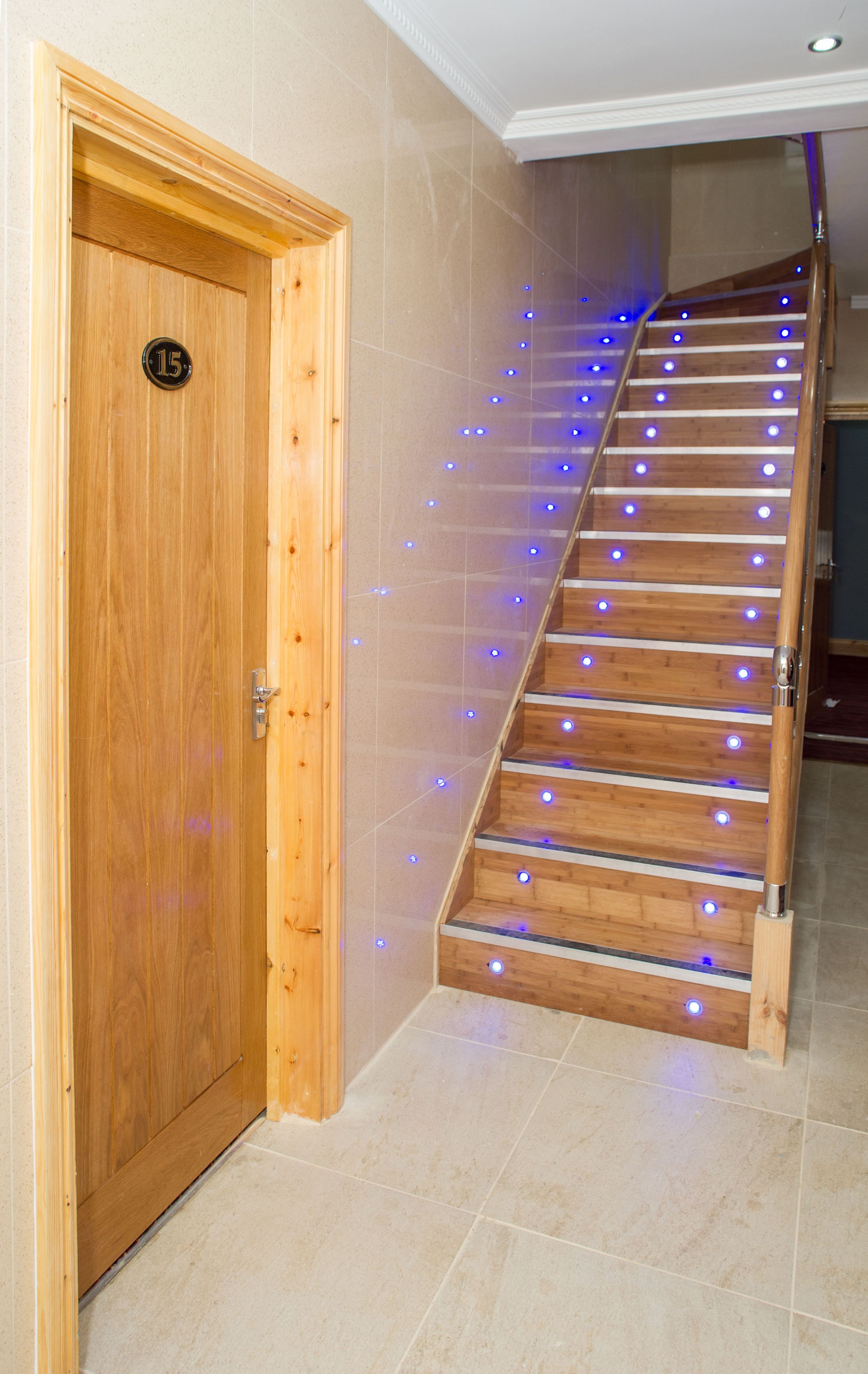 Una escalera de madera con luces LED azules, una puerta de madera con una placa de número y paredes con baldosas color beige.