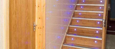 Una escalera de madera con luces LED azules, una puerta de madera con una placa de número y paredes con baldosas color beige.