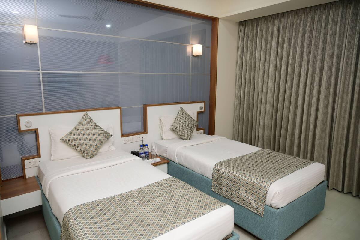 A hotel room with two beds, each with patterned bedspreads, a large window with blinds, and a curtain on the right.