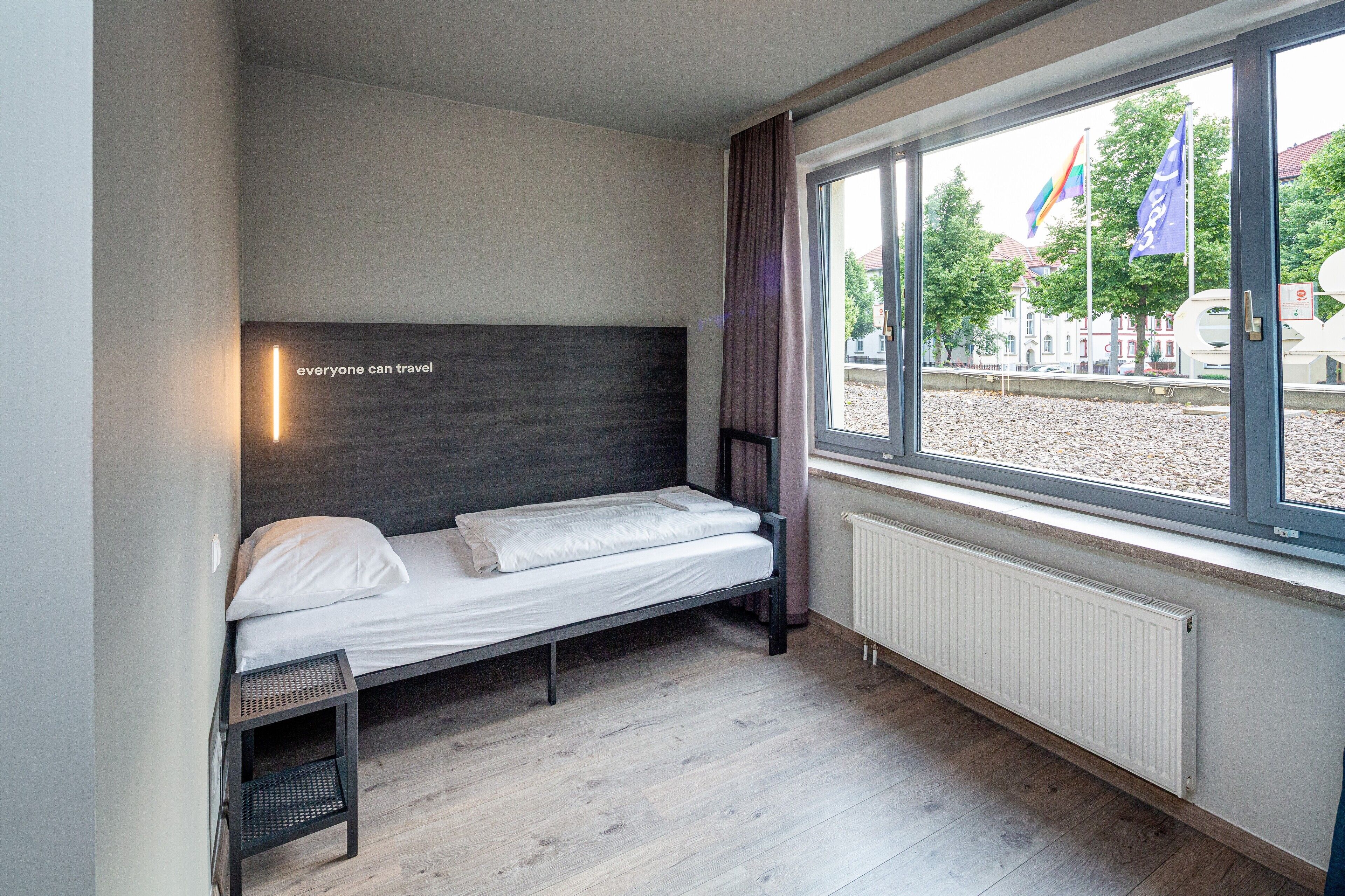 Una cama individual con cabecera, una mesita de noche pequeña, una ventana con cortinas y vistas al exterior.