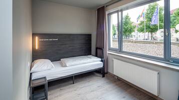 Una cama individual con cabecera, una mesita de noche pequeña, una ventana con cortinas y vistas al exterior.
