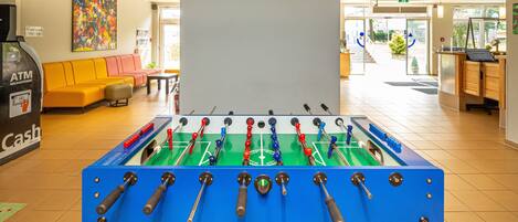 Une table de baby-foot dans un hall d’entrée bien éclairé, avec un canapé, un distributeur automatique de billets et un tableau accroché au mur.