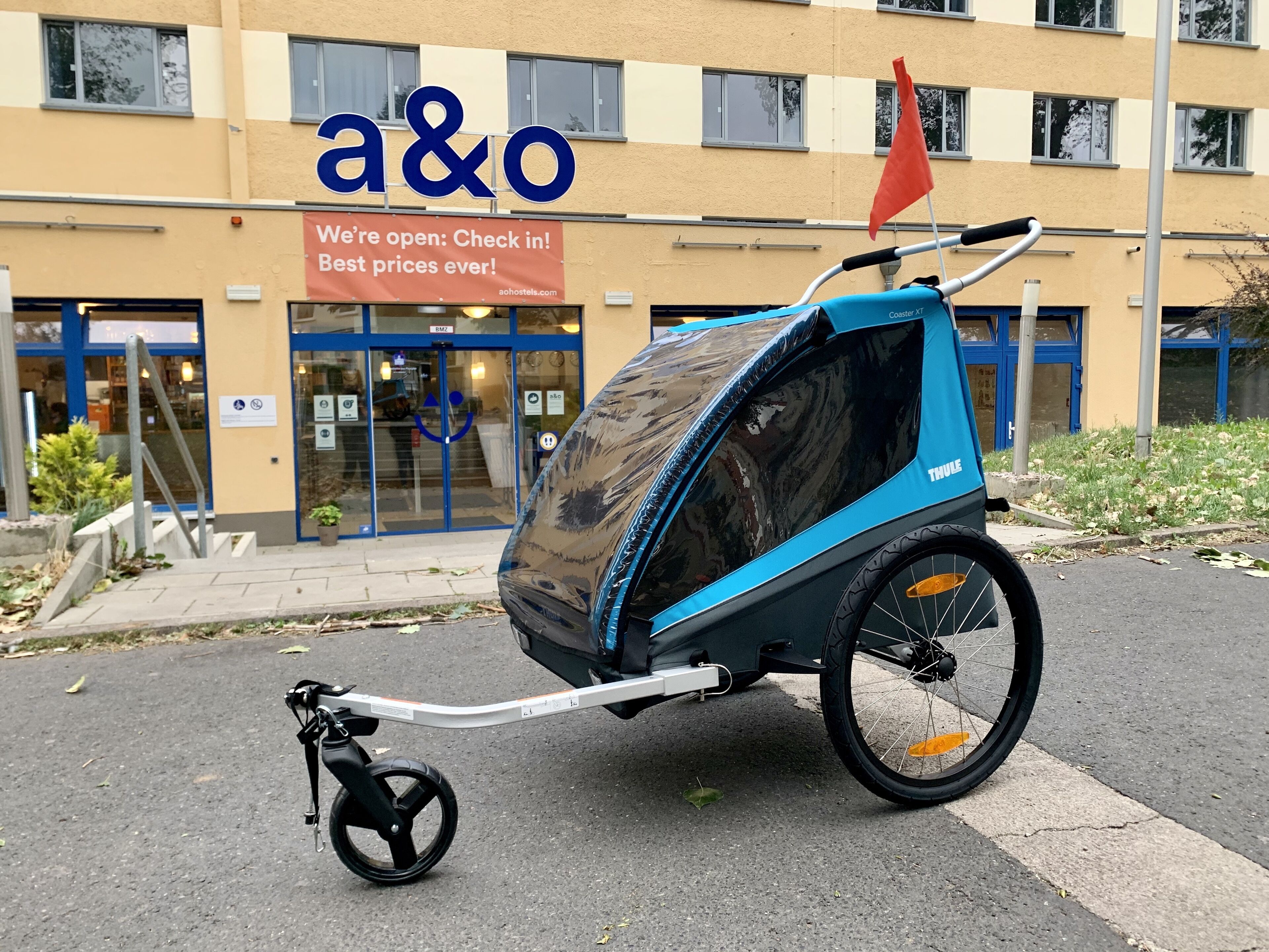 Ein blau-schwarzer Kinderwagen mit Verdeck, abgestellt vor einem Gebäude mit dem Schild "a&o Wir sind geöffnet: Einchecken! Beste Preise überhaupt!".