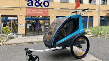 Ein blau-schwarzer Kinderwagen mit Verdeck, abgestellt vor einem Gebäude mit dem Schild "a&o Wir sind geöffnet: Einchecken! Beste Preise überhaupt!".