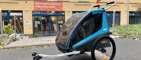 Un cochecito azul y negro con dosel, estacionado frente a un edificio con un letrero que dice "a&o We're open: Check in! Best prices ever!"