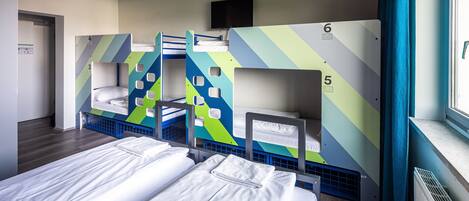 A dormitory room with bunk beds numbered 3 to 6, a television above the beds, and a window with curtains.
