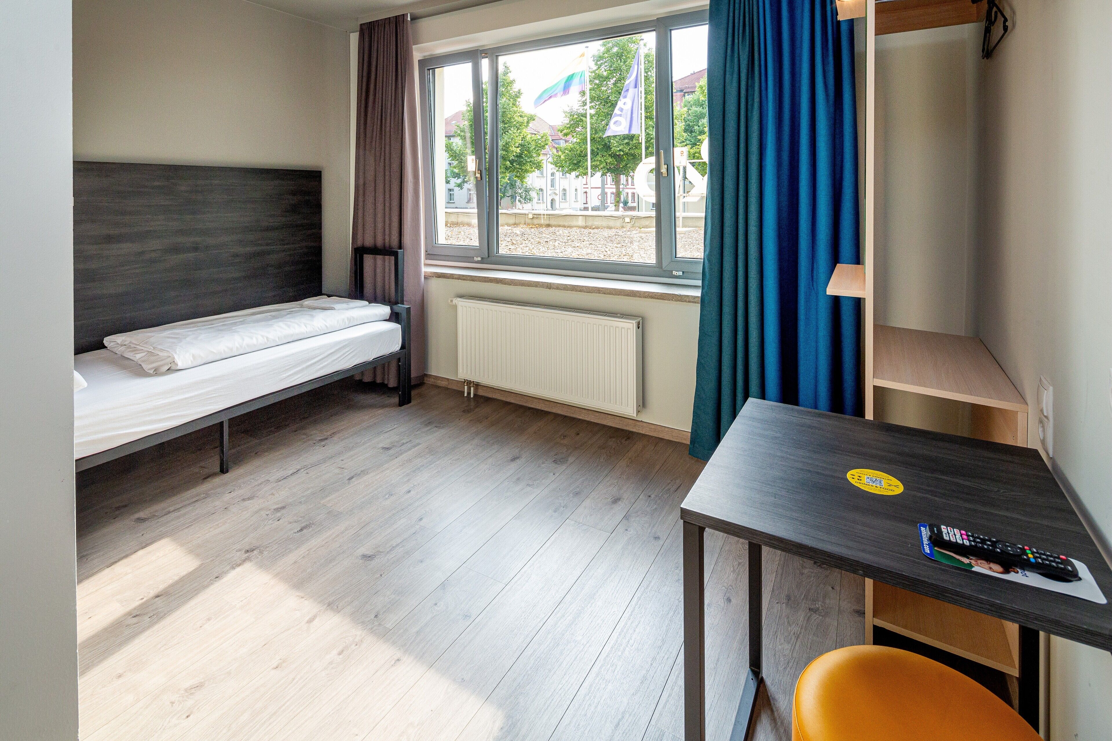 Una habitación pequeña con una cama individual, una mesa y un control remoto, y un ventanal con cortinas.