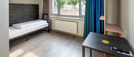Una habitación pequeña con una cama individual, una mesa y un control remoto, y un ventanal con cortinas.