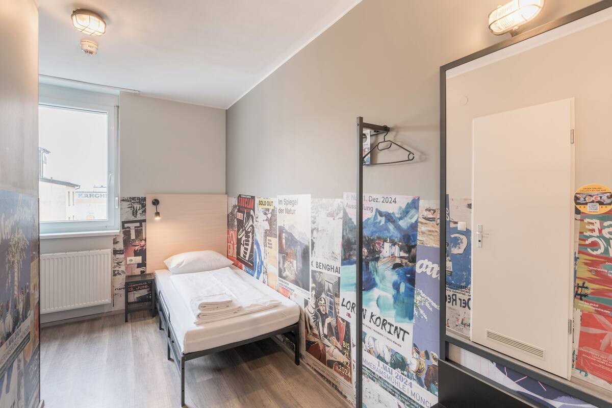 Un lit simple dans une chambre avec une fenêtre, des affiches au mur et une porte.