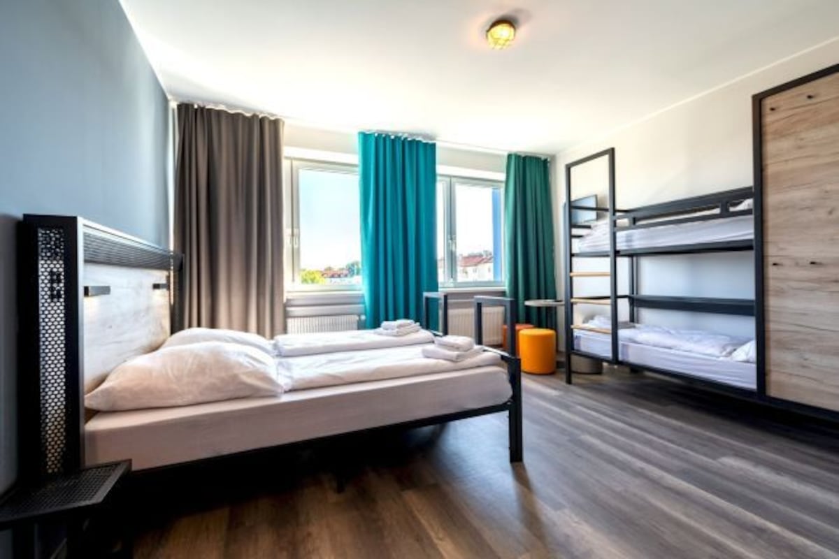 Une chambre d’hôtel moderne équipée d’un lit simple, d’un lit superposé, d’un bureau avec un ordinateur portable et d’une fenêtre avec des rideaux.