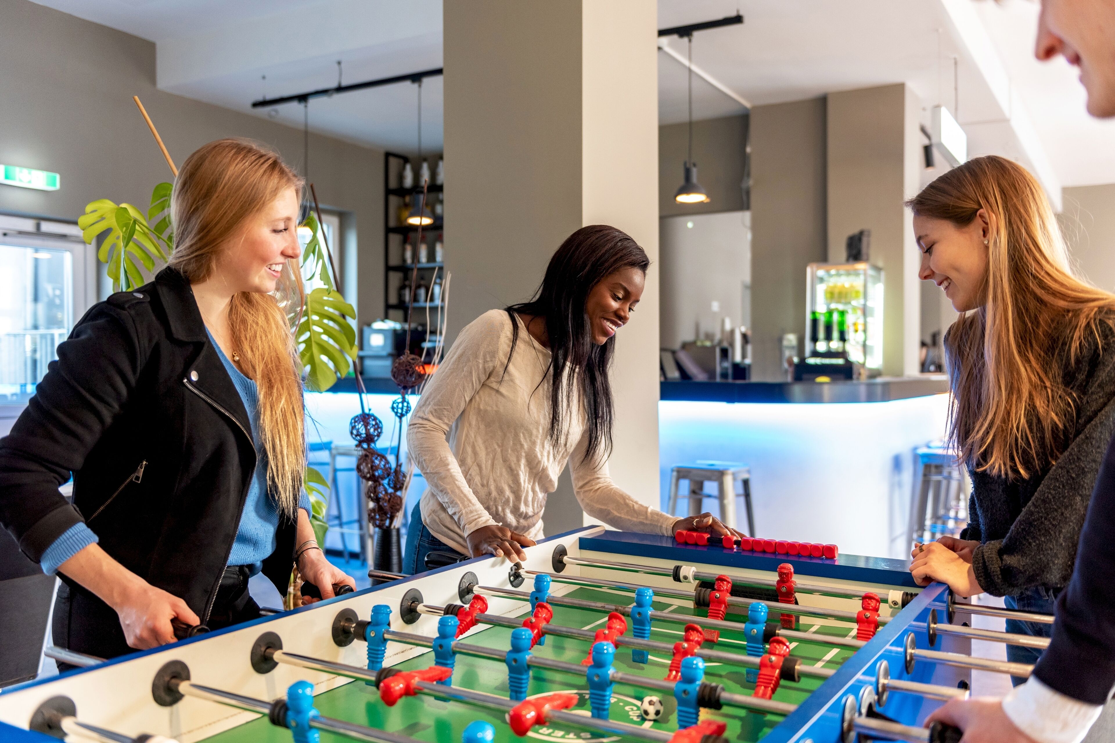 Eine Gruppe von Personen spielt Kicker in einem modernen Innenbereich, im Hintergrund ist eine Bar zu sehen.