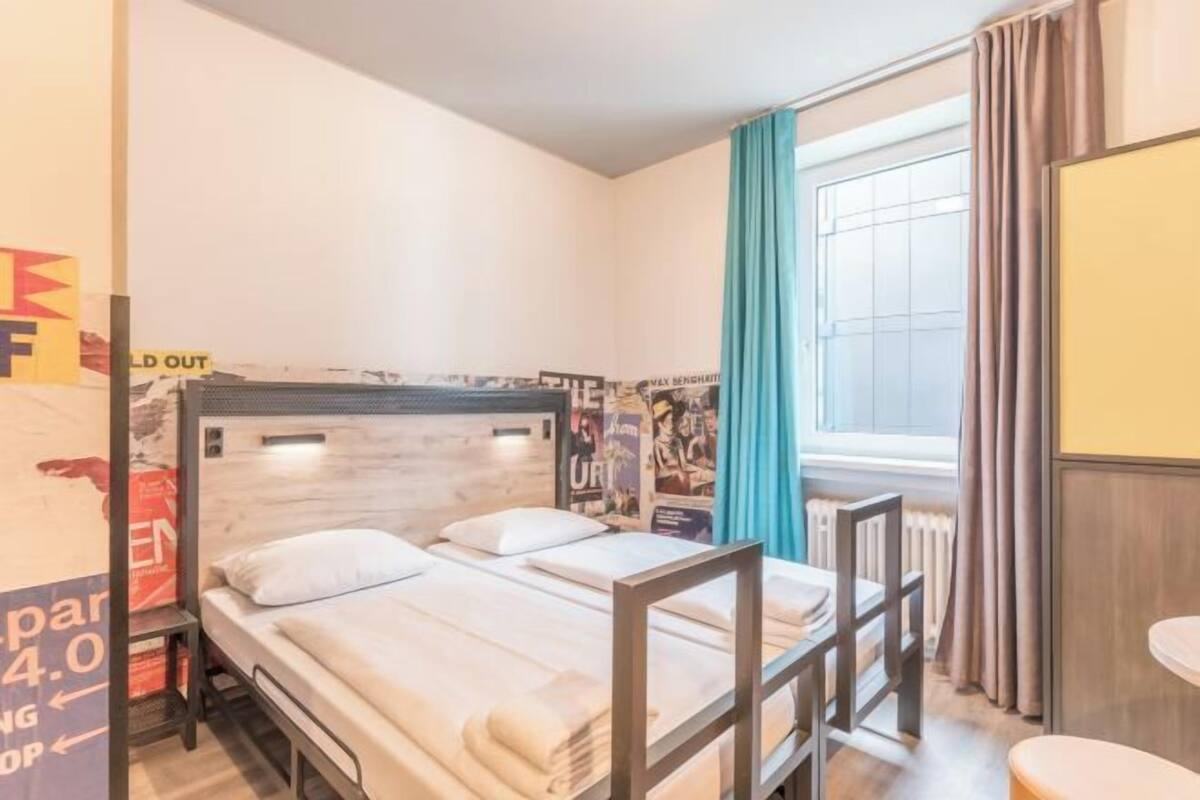 Une chambre avec un lit simple, une tête de lit en bois, une petite table, une fenêtre avec des rideaux et un radiateur.