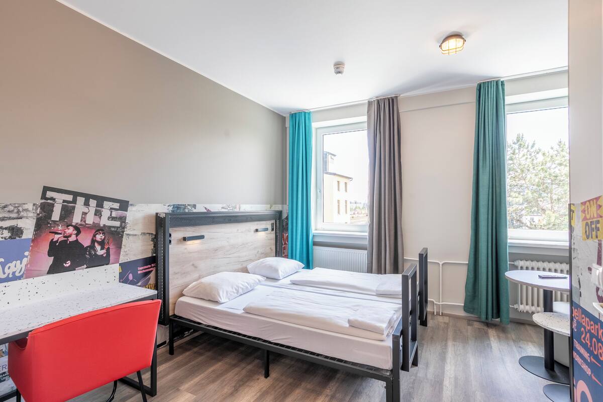 Une chambre d’hôtel avec un lit, un bureau, une chaise, une fenêtre avec des rideaux et une affiche au mur.