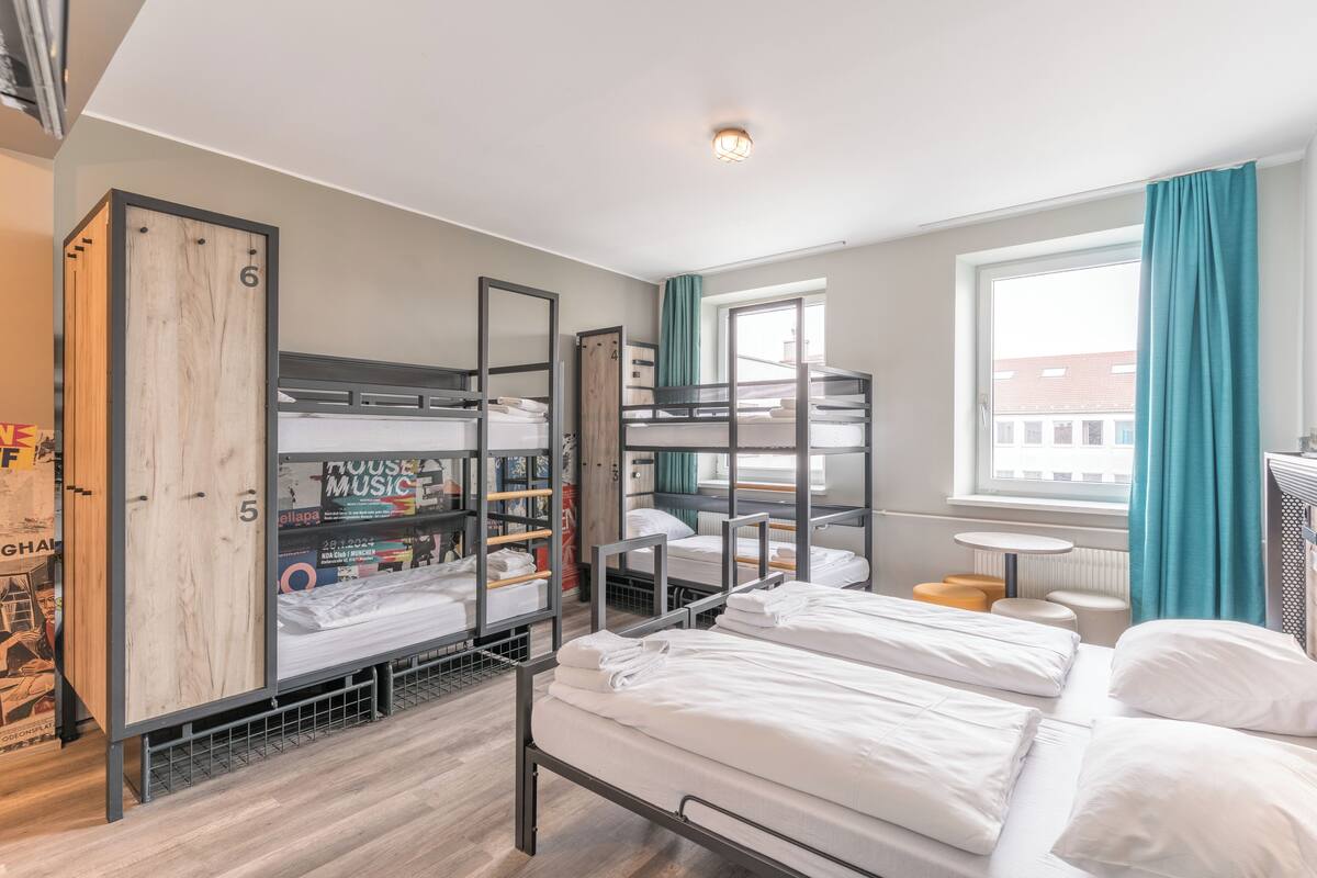 Une chambre de type dortoir, avec des lits superposés, une petite table et une fenêtre avec des rideaux.