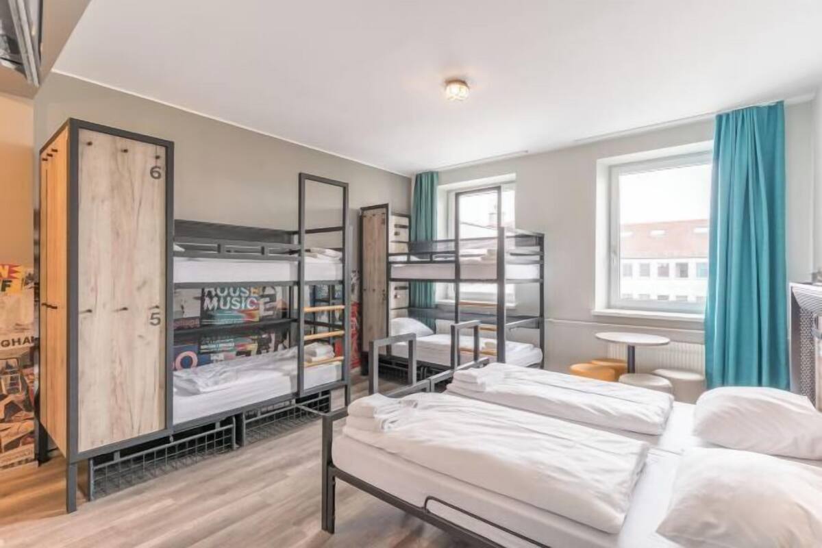 Une chambre de type dortoir, avec des lits superposés, une petite table ronde et une fenêtre avec des rideaux.