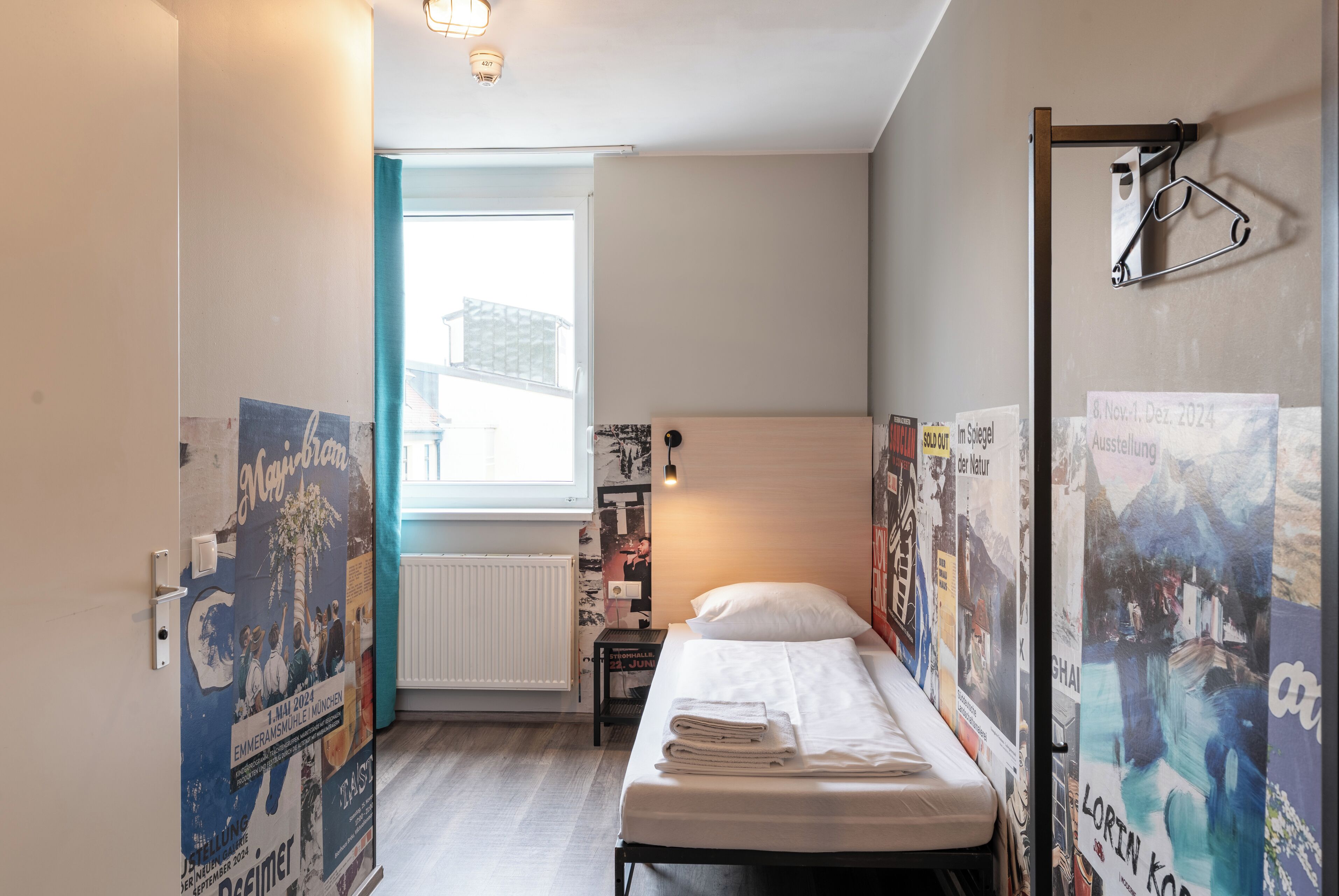 A small, well-lit room with a single bed, a radiator, and various posters on the walls.