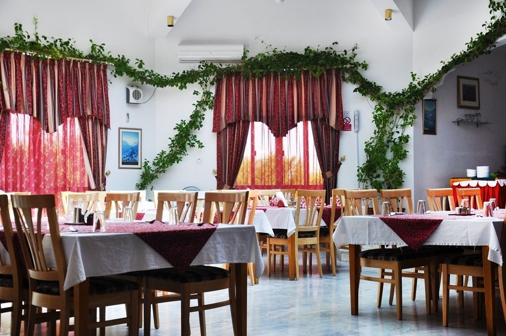 Un espace repas avec des tables dressées pour le service, des chaises en bois et des rideaux rouges ornés de motifs de lierre vert.