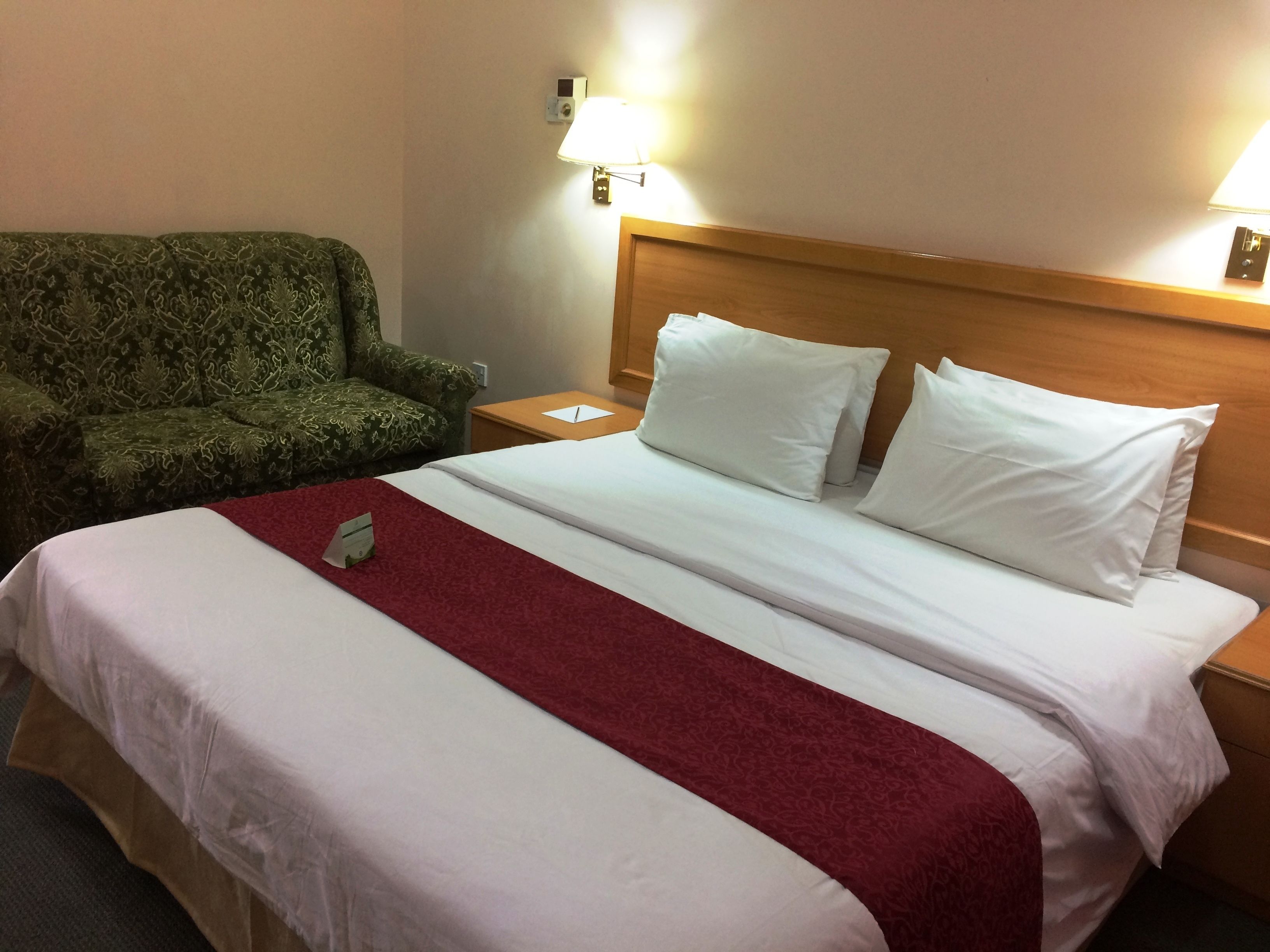Une chambre d’hôtel avec un grand lit, un fauteuil vert, une table de chevet avec une lampe et une tête de lit en bois.