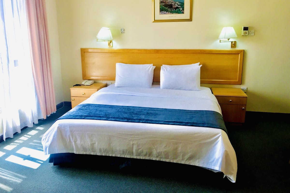 A hotel room with a large bed, two bedside tables, a nightstand, a lamp, a painting on the wall, and a window with curtains.