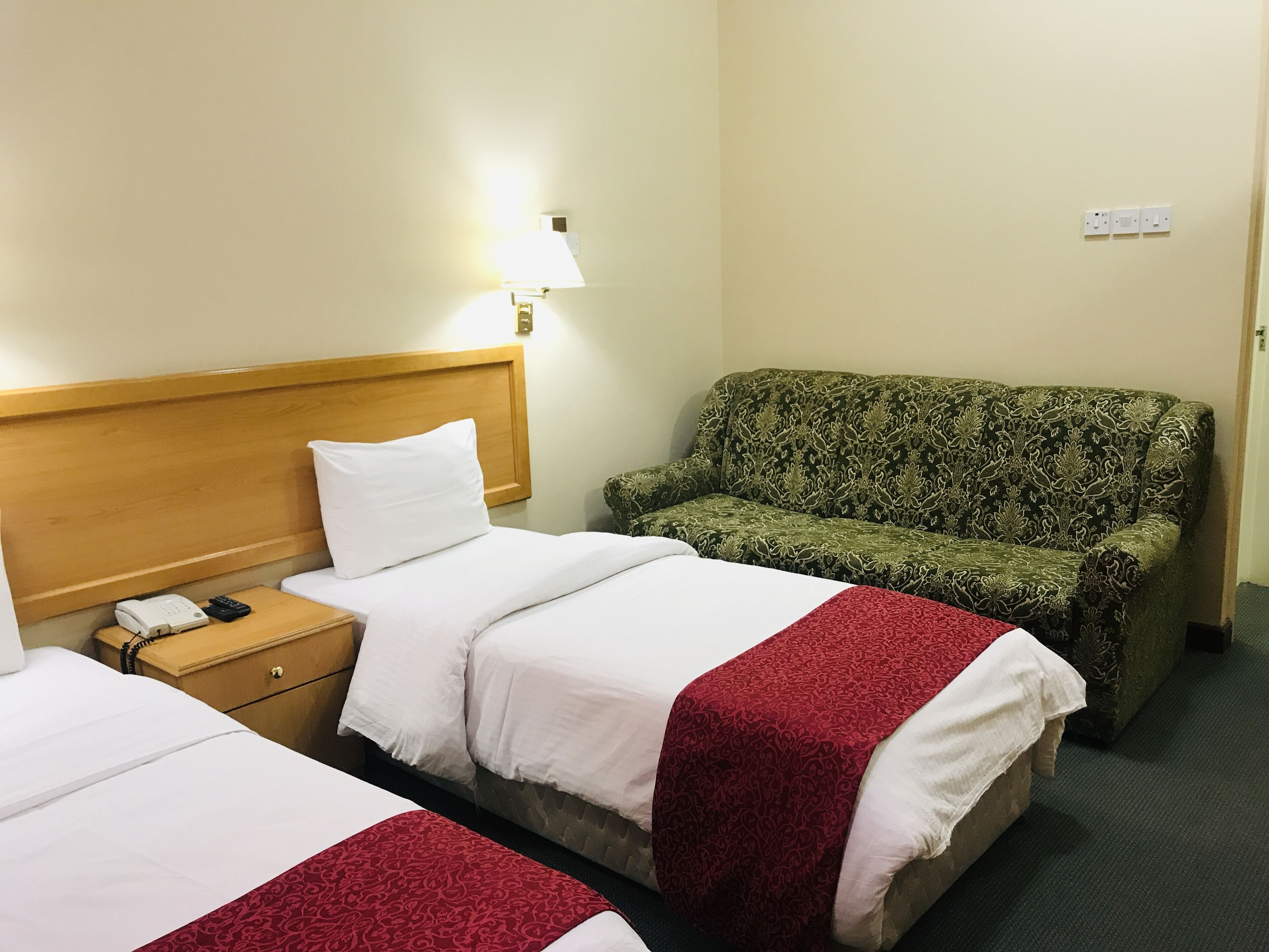 Una camera d'albergo con due letti, una testiera in legno, un divano verde con motivi decorativi e una lampada da comodino.