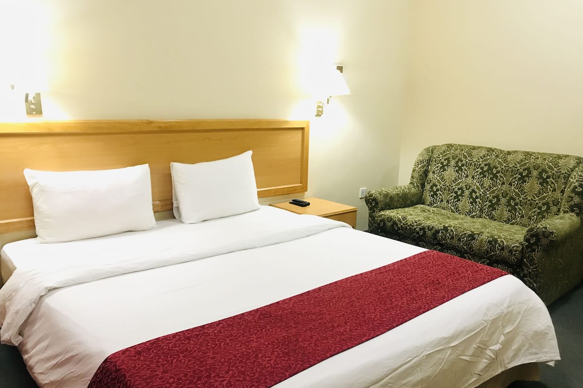 A hotel room with a bed, two pillows, a nightstand, a remote control, and a green patterned armchair.