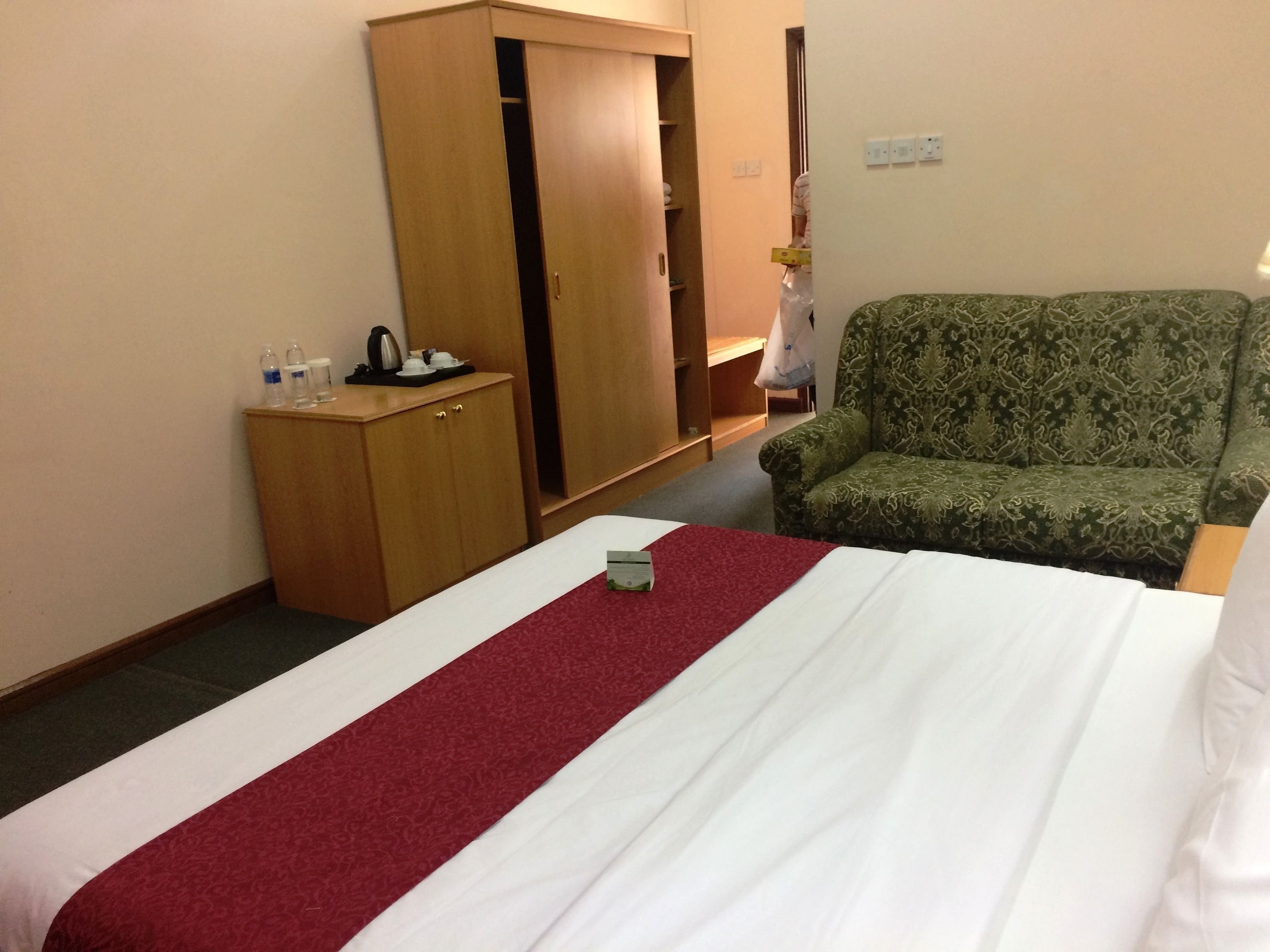 A hotel room with a bed, a green patterned sofa, a wooden wardrobe, and a small table with a kettle and cups.