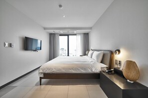 A modern hotel room with a large bed, a flat-screen TV mounted on the wall, a bedside table with a lamp, and a view of the city through the window. - Lareem Boutique Hotel (Seogwipo)