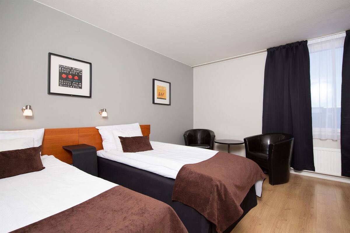 Ein Hotelzimmer mit zwei Betten, einem kleinen Tisch, einem Stuhl und einem Fenster mit Vorhängen.
