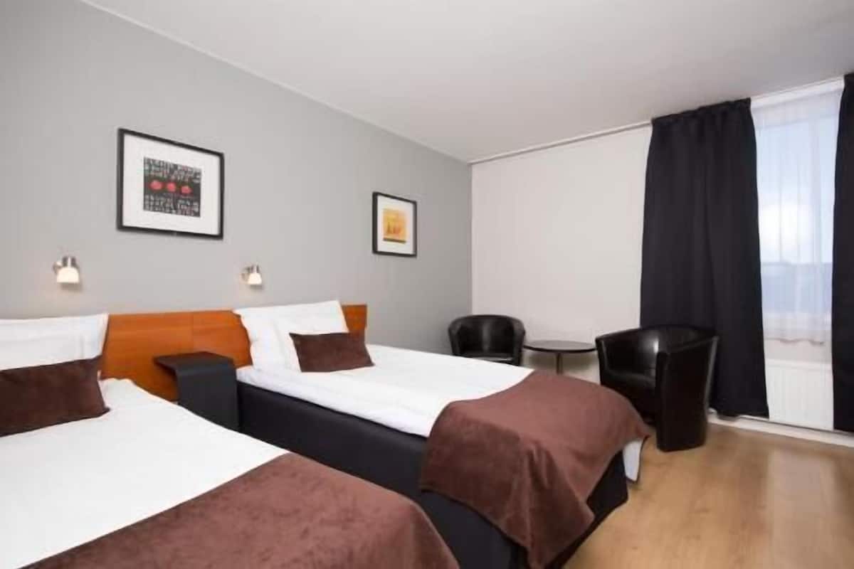 Ein Hotelzimmer mit zwei Betten, einem kleinen Tisch, einem Stuhl und einem Fenster mit Vorhängen.