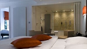 Habitación de hotel moderna con una cama grande, dos almohadas naranjas, un ordenador portátil sobre una mesita auxiliar, zona de ducha y vistas al exterior.