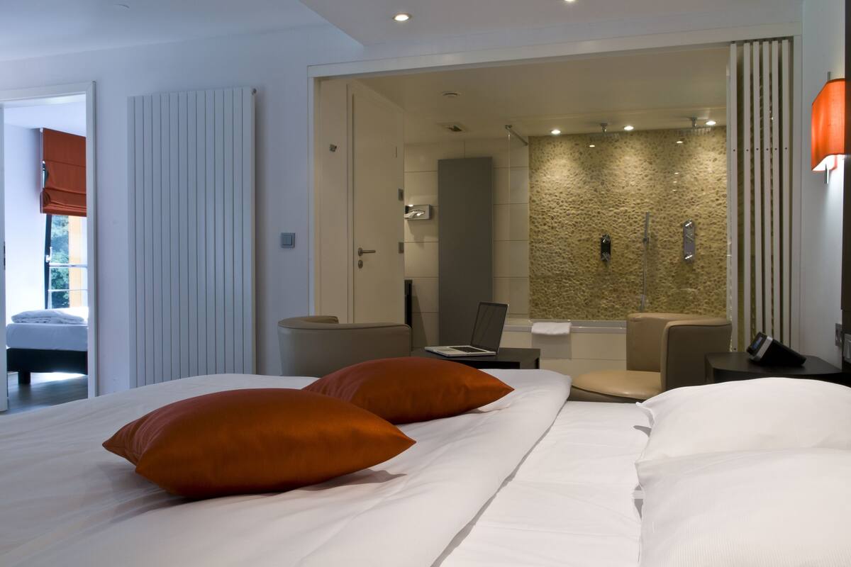 A modern hotel room with a large bed, two orange pillows, a laptop on a side table, a shower area, and a view of the outside.