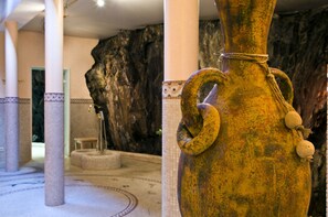A large, rustic vase with a rope handle, set against a backdrop of a stone wall and columns in a lobby area.
