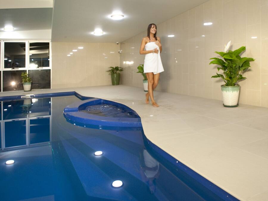 A woman in a white dress standing by a pool with a hot tub, surrounded by potted plants and a tiled wall.
