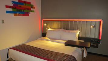 A modern hotel room with a neatly made bed, a headboard with integrated lighting, a bedside table, and a wall art piece.