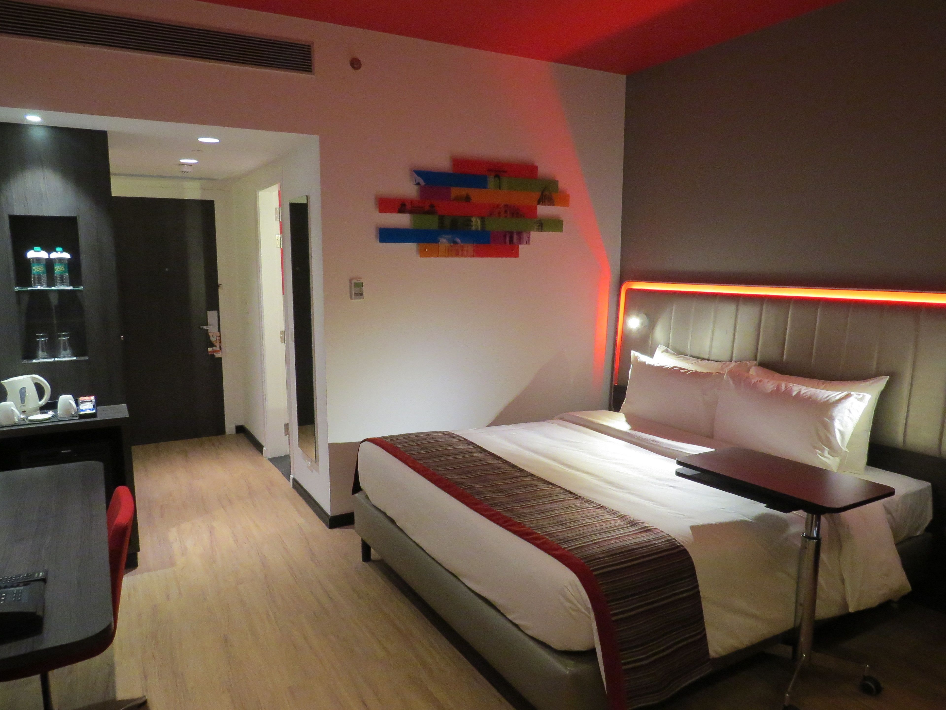 Una habitación de hotel moderna con cama, mesita de noche y escritorio con silla. Hay un espejo en la pared y una puerta que da a otra habitación.