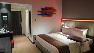 Una habitación de hotel moderna con cama, mesita de noche y escritorio con silla. Hay un espejo en la pared y una puerta que da a otra habitación.