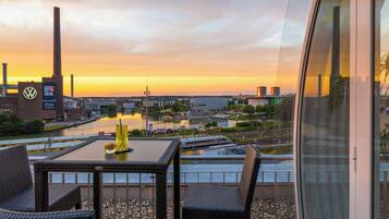 A terrace with wicker furniture, a table, and a view of a river, buildings, and a bridge during sunset.